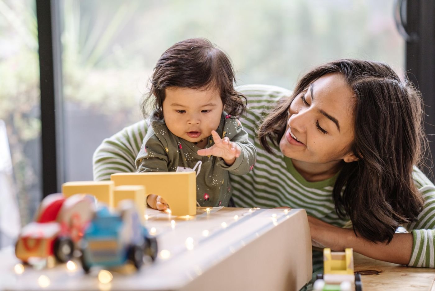 A mother smiles as her toddler reaches for a wooden block toy